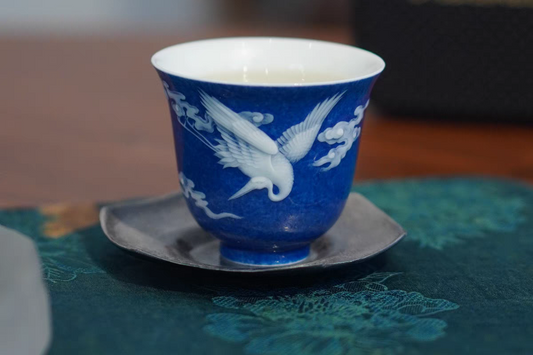 A blue and white ceramic cup with a bird pattern, placed on a saucer, on a floral patterned surface.